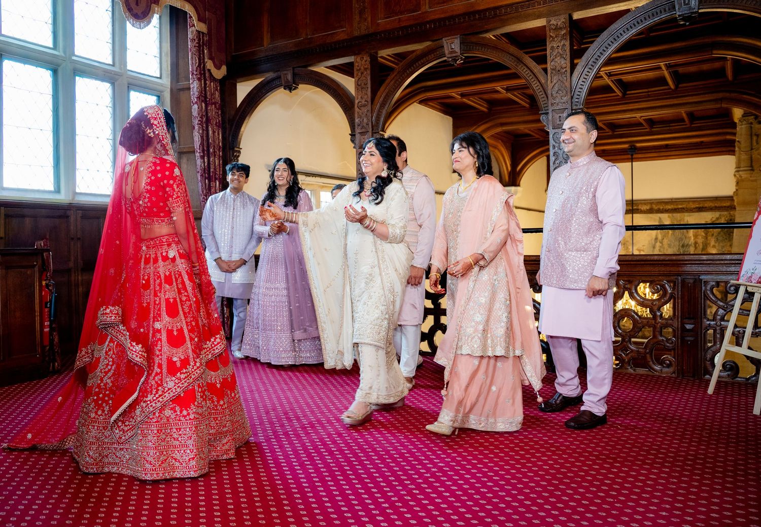 Indian bride in red lehenga greeting family in ornate wedding venue hall.