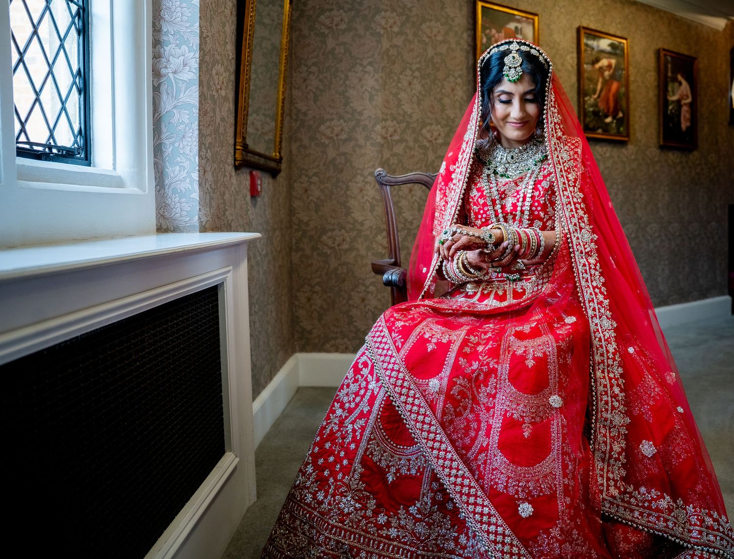 Beautiful Indian bride in red embroidered lehenga with dupatta seated near window in elegant room.