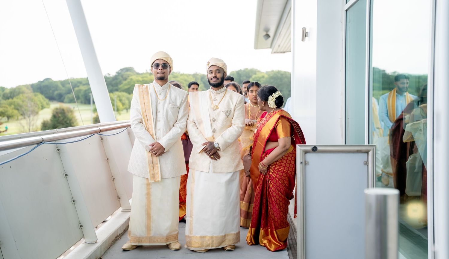 Two grooms in white and gold traditional Indian wedding attire stand on a balcony with family members.