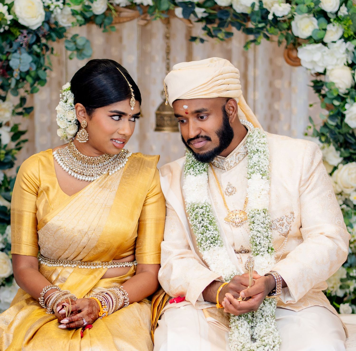 South Indian bride in gold silk saree and groom in cream sherwani share an intimate moment at their wedding ceremony.