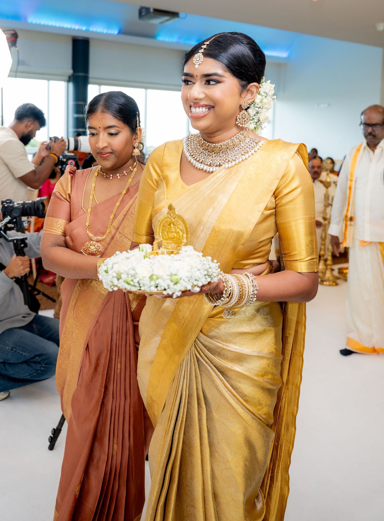 Two women in traditional golden and bronze silk sarees with jewelry holding flowers at a cultural ceremony.