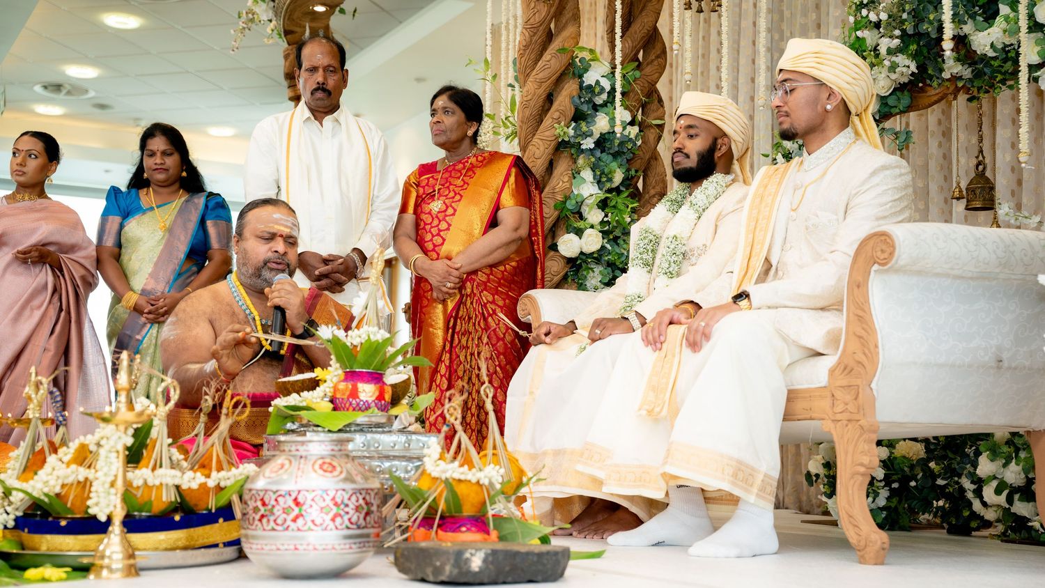 A diverse group of people in traditional and ceremonial attire gathered around a table with fruits and flowers at an indoor celebration.