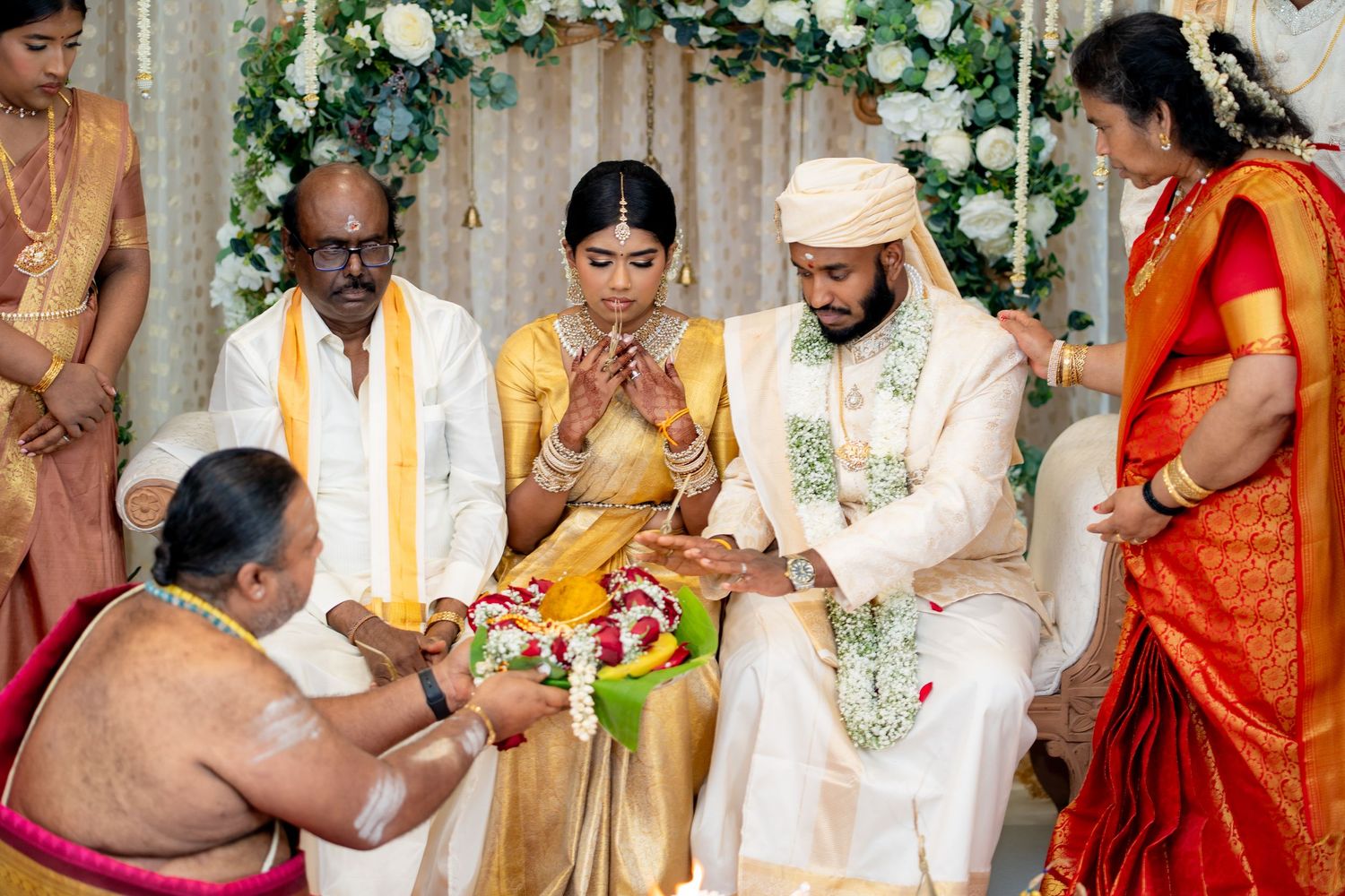 Traditional South Indian wedding ceremony with bride in gold saree and groom in white sherwani exchanging vows with priest.