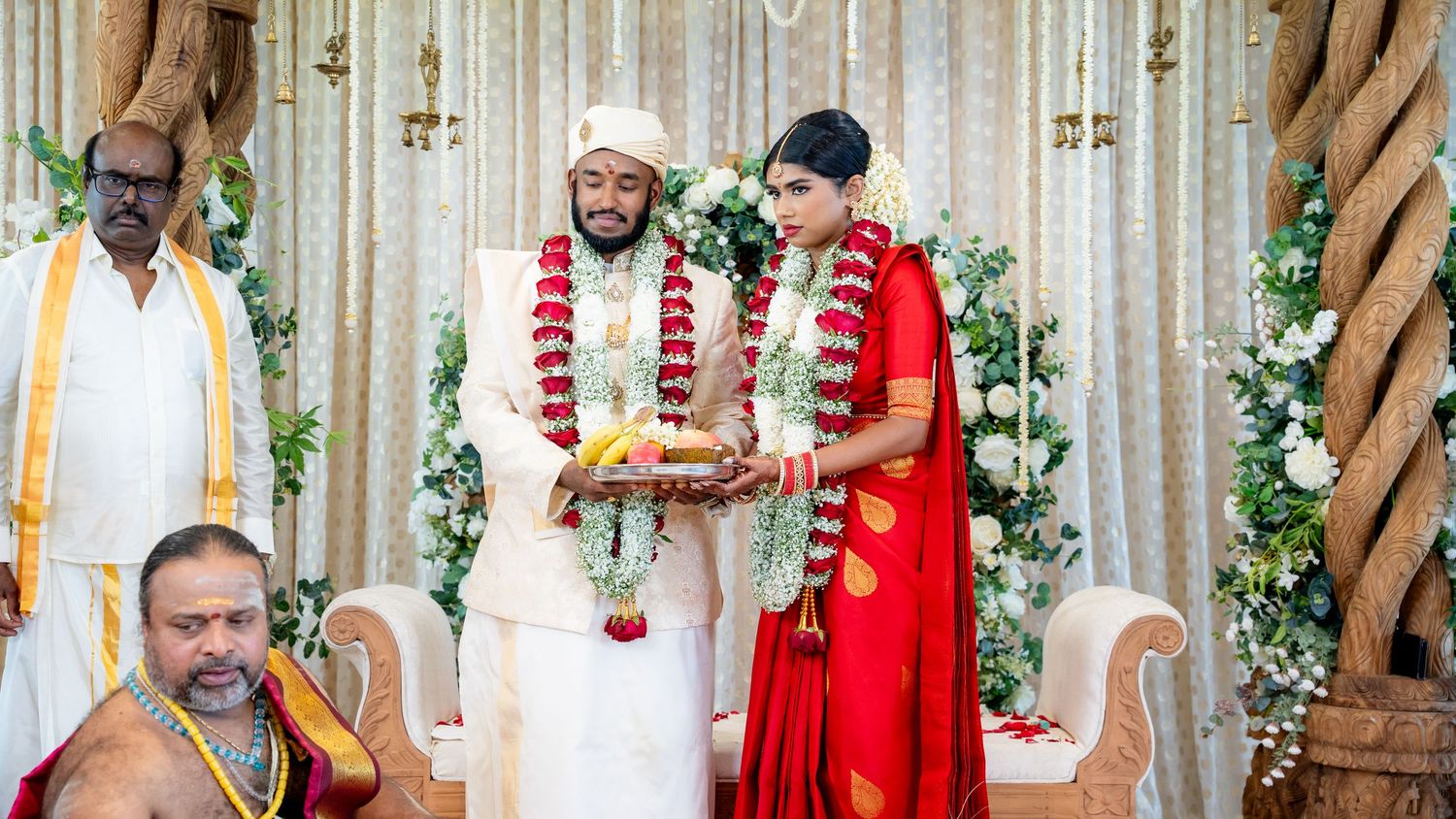 South Indian newlyweds holding ceremonial plate together on decorated wedding mandap with priest and family members.