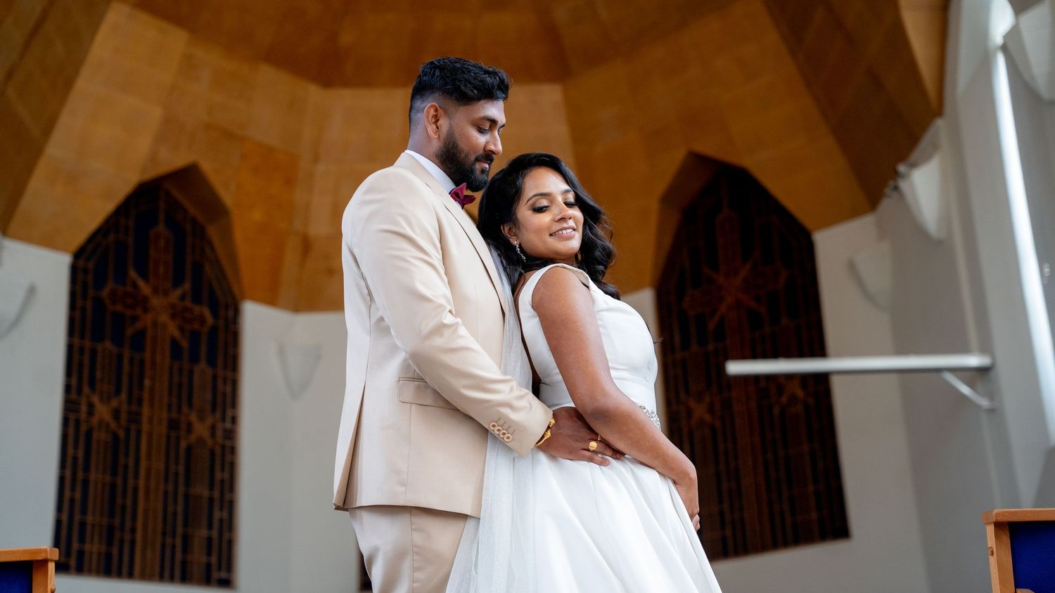 A couple in wedding attire poses elegantly inside a chapel, groom in beige suit, bride in white gown.
