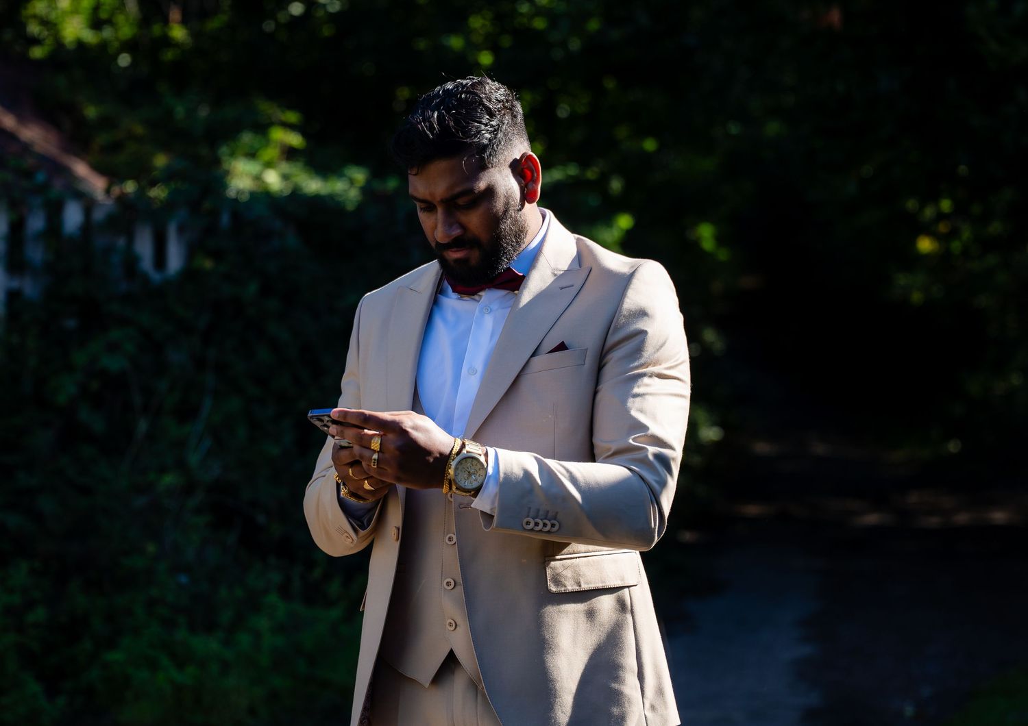 Groom in beige three-piece suit checks phone outdoors in dappled sunlight at wedding venue.