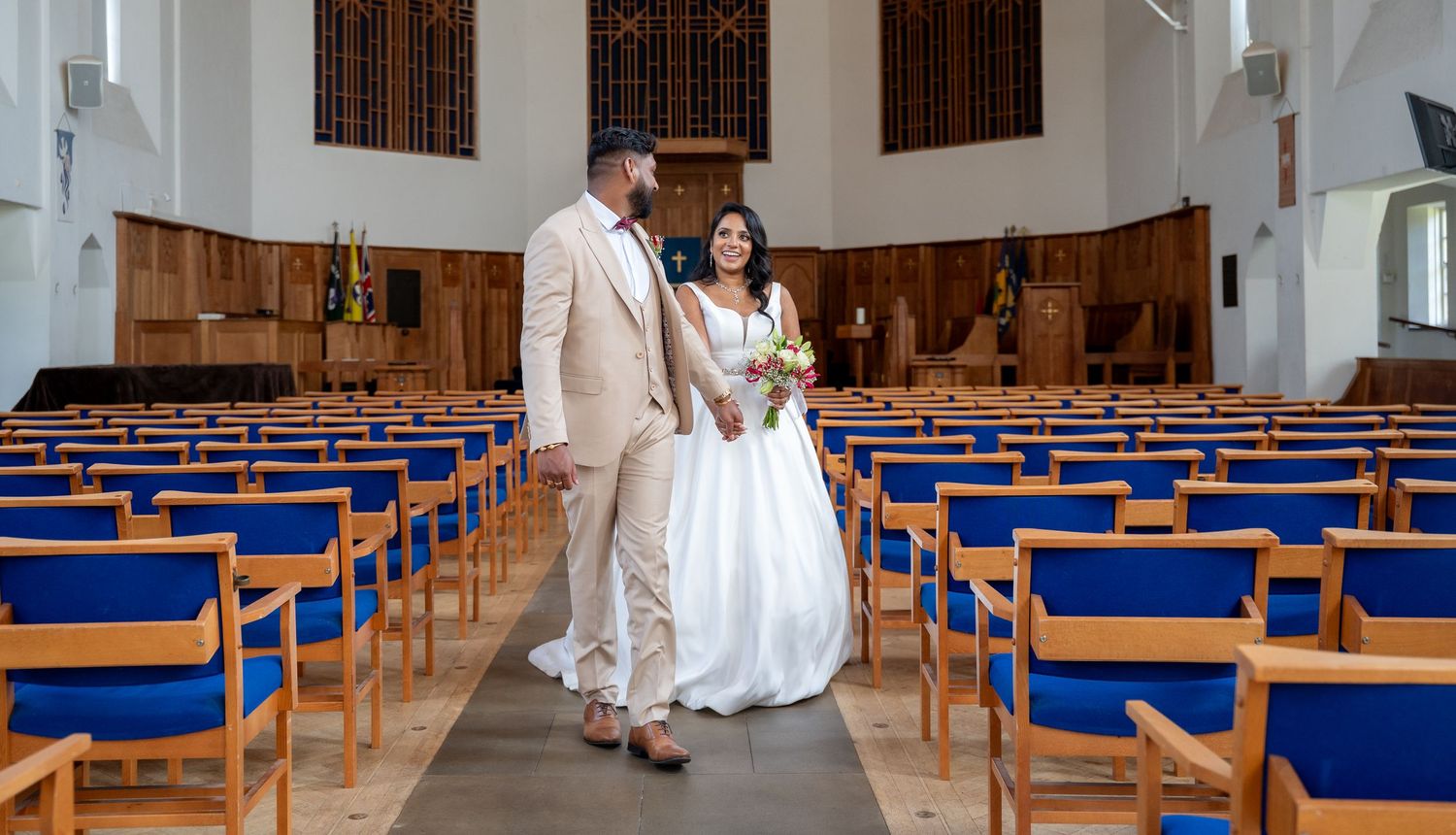 Bride and groom walk together down a church aisle lined with blue-cushioned wooden pews after their ceremony.