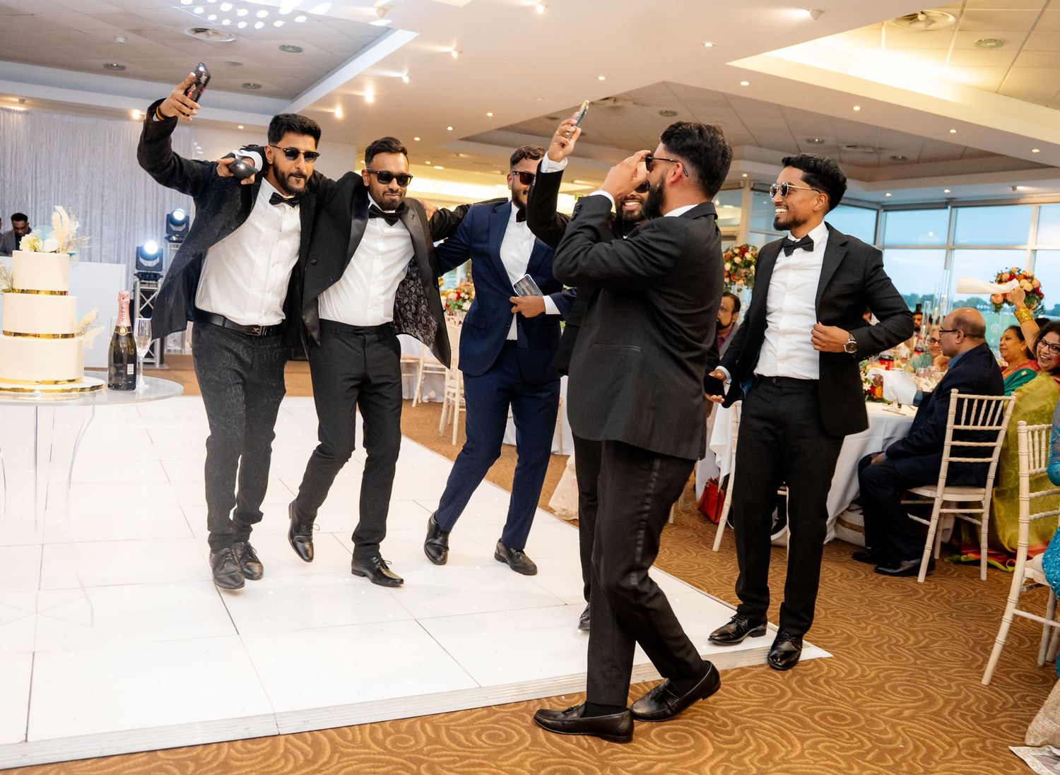 Groomsmen in black suits and sunglasses dancing and taking selfies on the wedding dance floor.