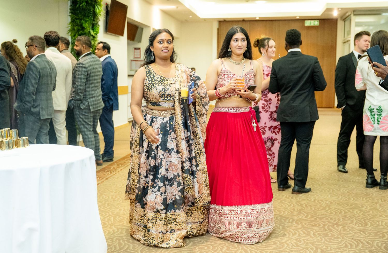 Two women in vibrant Indian lehenga outfits posing together at an elegant formal event.