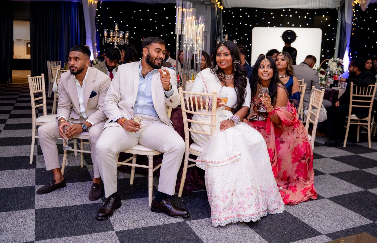 Guests in formal attire smile and relax on white chairs at an elegant event with a starlit backdrop.