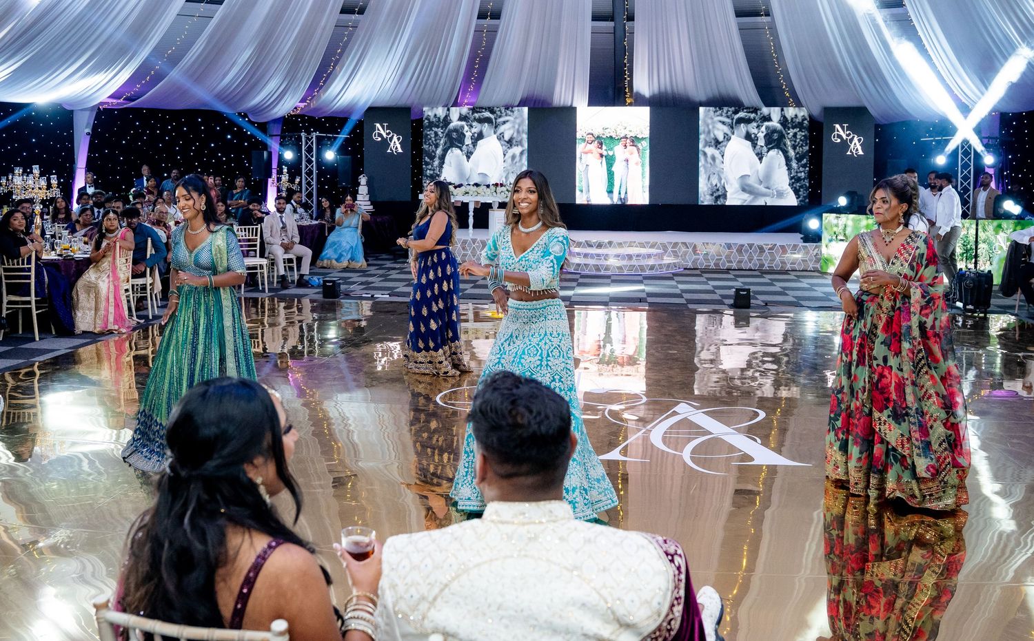 Women in colorful Indian attire performing a dance for bride and groom at wedding reception.