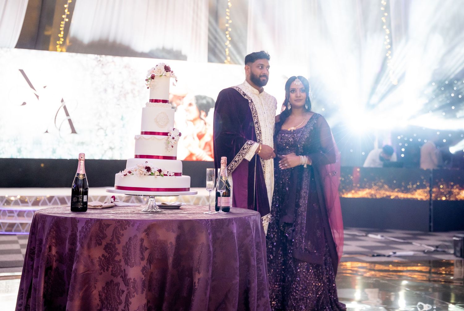 Bride and groom in purple Indian wedding attire standing beside elegant white wedding cake.