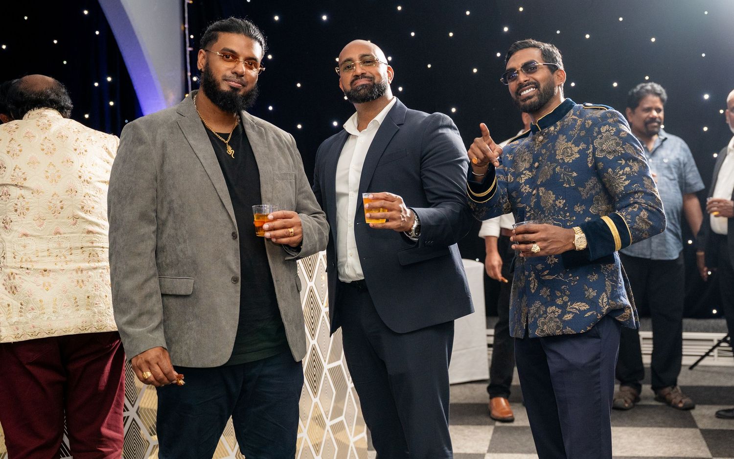 Three well-dressed men smiling and holding drinks at an elegant evening event with a sparkling backdrop.