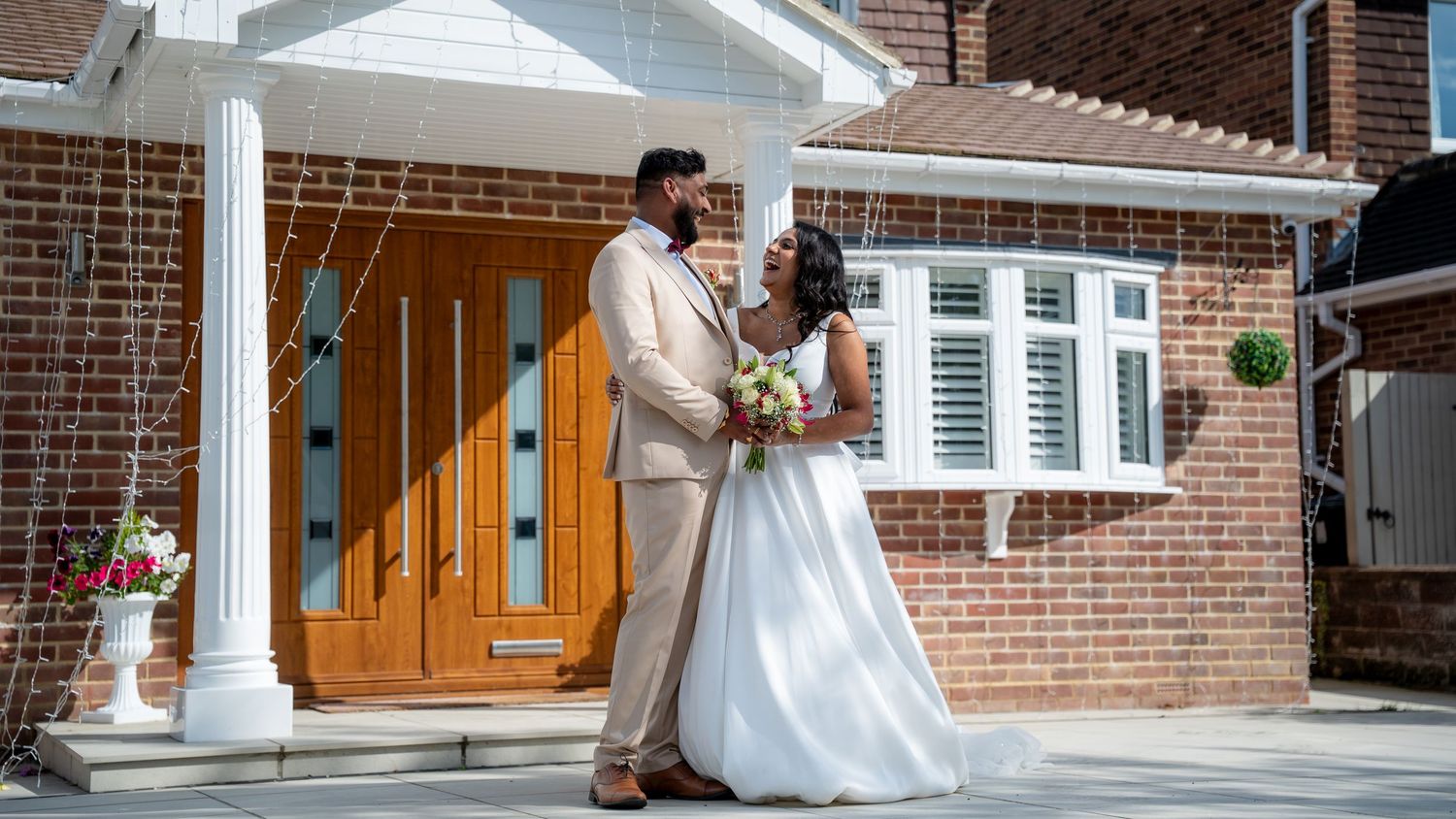 Bride in white gown and groom in tan suit pose together outside a brick house with a wooden door.