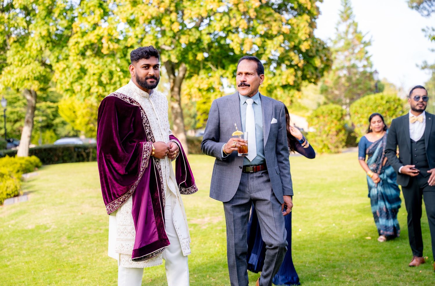 A groom in a purple velvet sherwani walks with an older man in a suit at an outdoor wedding.