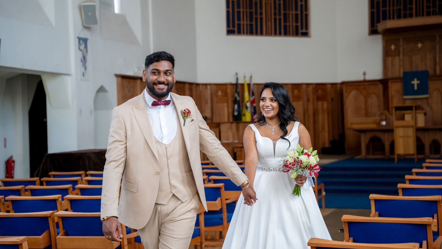 Smiling bride and groom holding hands walking down church aisle, groom in tan suit, bride in white gown with bouquet.