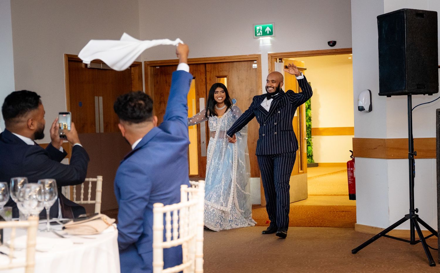 Groom raises white cloth welcoming bride in blue lehenga at elegant indoor wedding reception entrance.