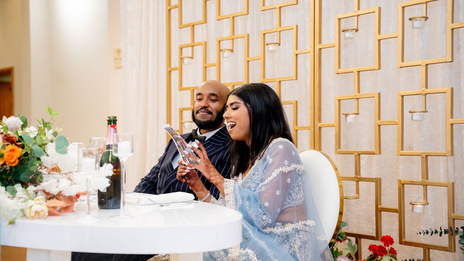 A bride in a blue embellished outfit and groom in a pinstripe suit share a moment at their wedding table.