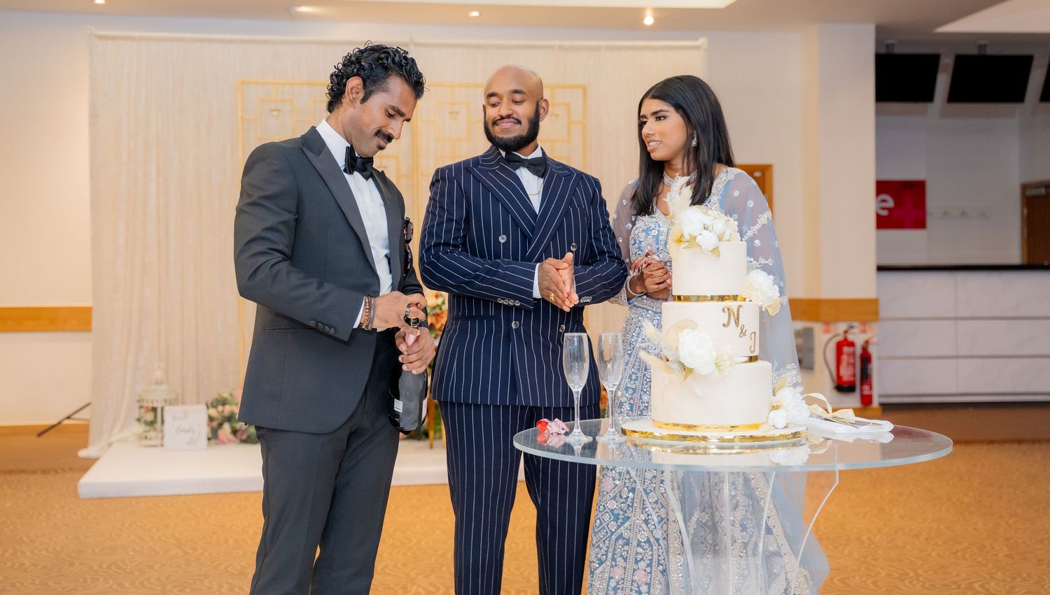 Two men and a bride posing with champagne beside a white wedding cake at a reception.