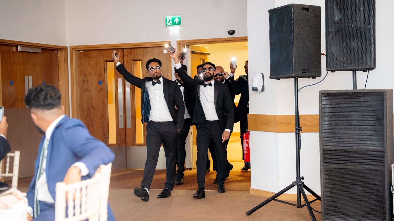 Three groomsmen in black tuxedos with bow ties entering a wedding venue with hands raised in celebration.