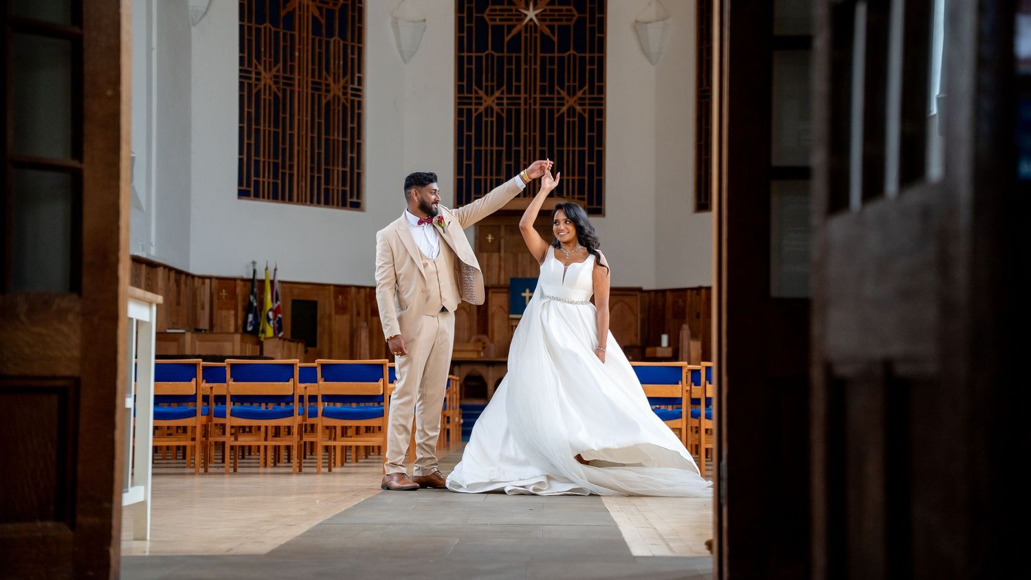 Bride and groom dancing elegantly in a sunlit church, groom in beige suit twirling bride in white wedding gown.