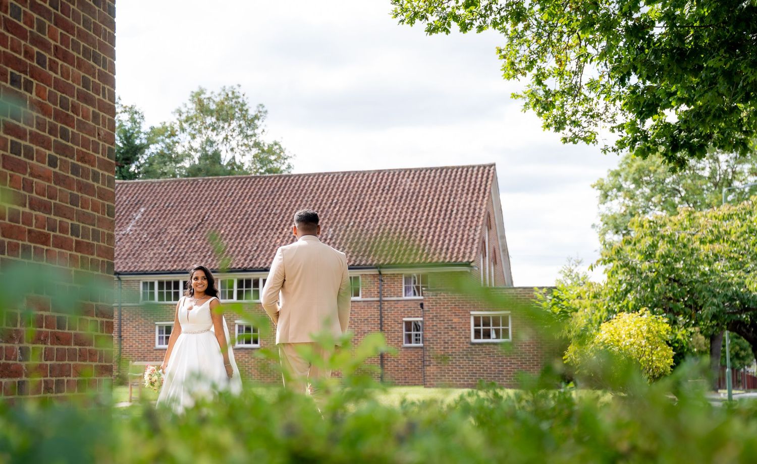 Bride and groom walking hand in hand across lush green gardens beside a charming brick building on their wedding day.