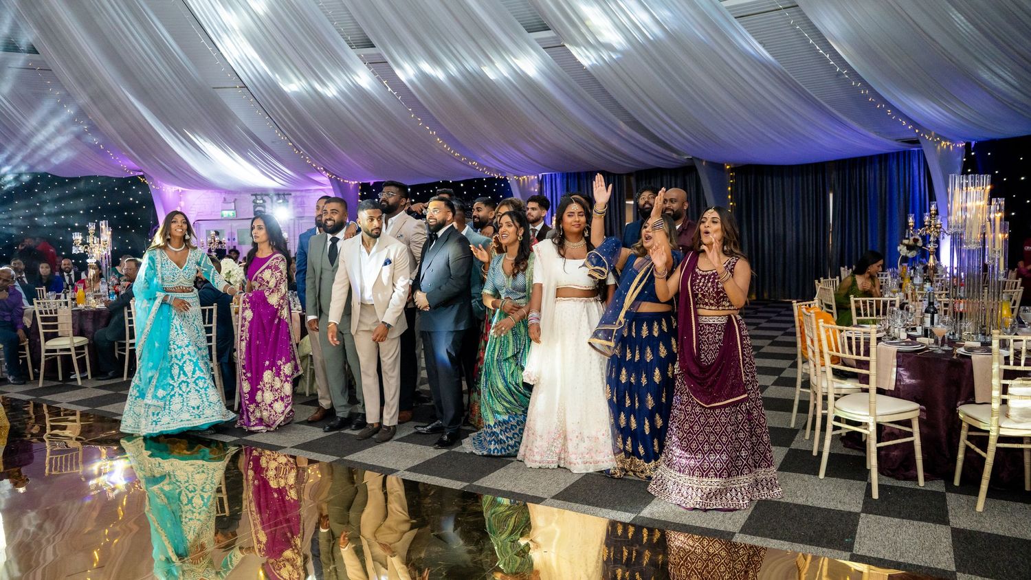 Group of guests in colorful traditional Indian attire celebrating on a dance floor at a wedding.