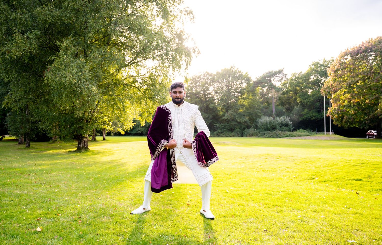 Groom in white sherwani and burgundy velvet dupatta posing confidently in sunlit green garden.