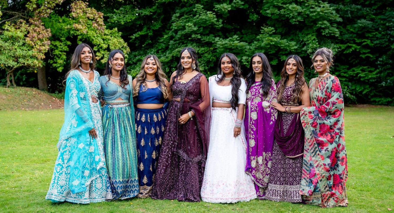 Eight women in vibrant Indian traditional attire, including lehengas and sarees, posing together outdoors at a wedding celebration.