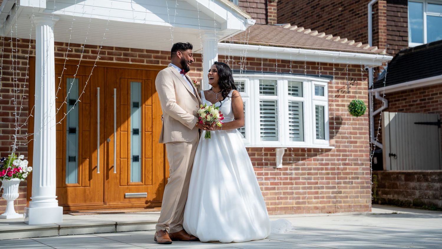 Elegant bride and groom posing together outside a brick home on their wedding day, wearing white gown and beige suit.