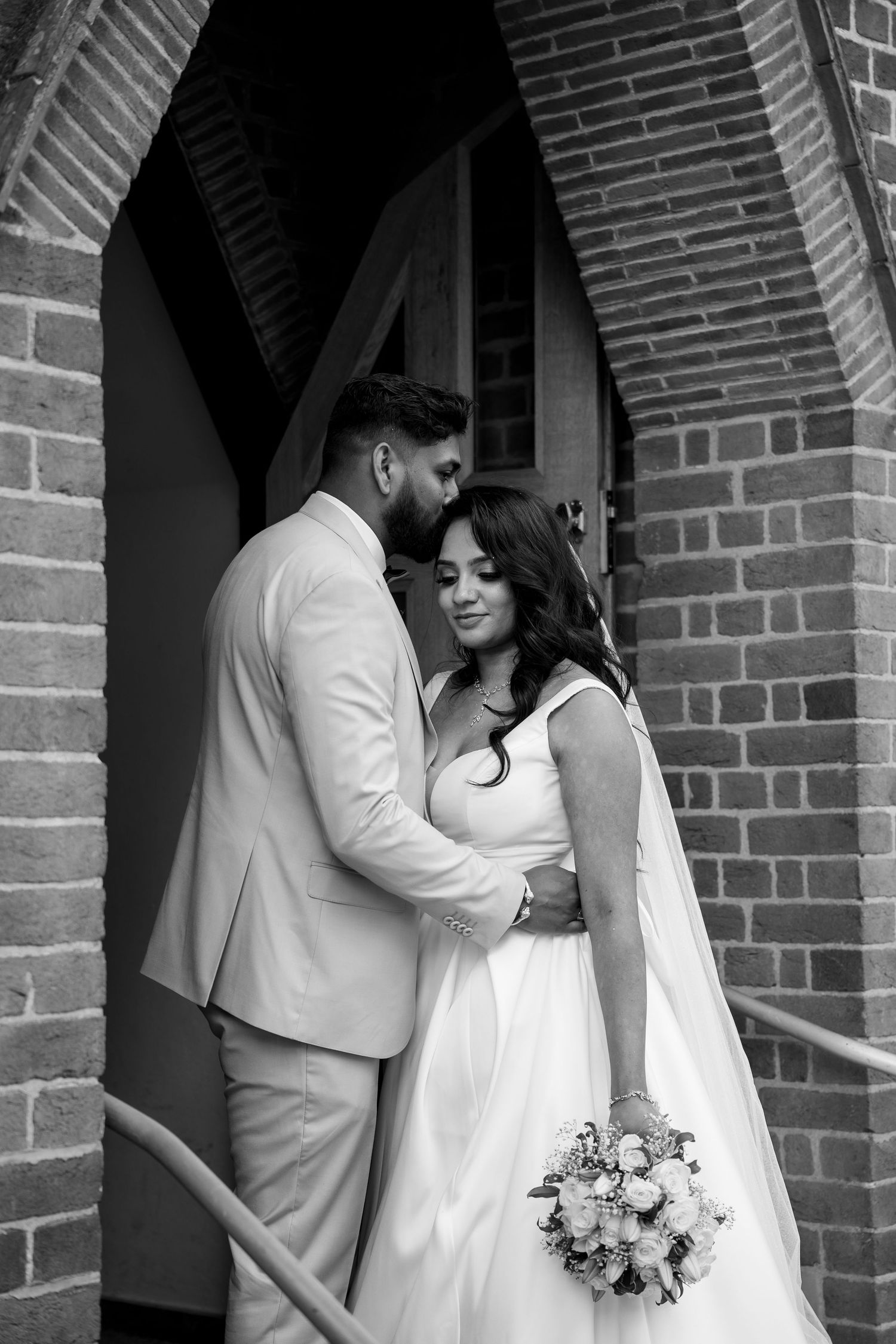 Black and white photo of a bride and groom embracing in a brick church doorway, bride holding a bouquet.