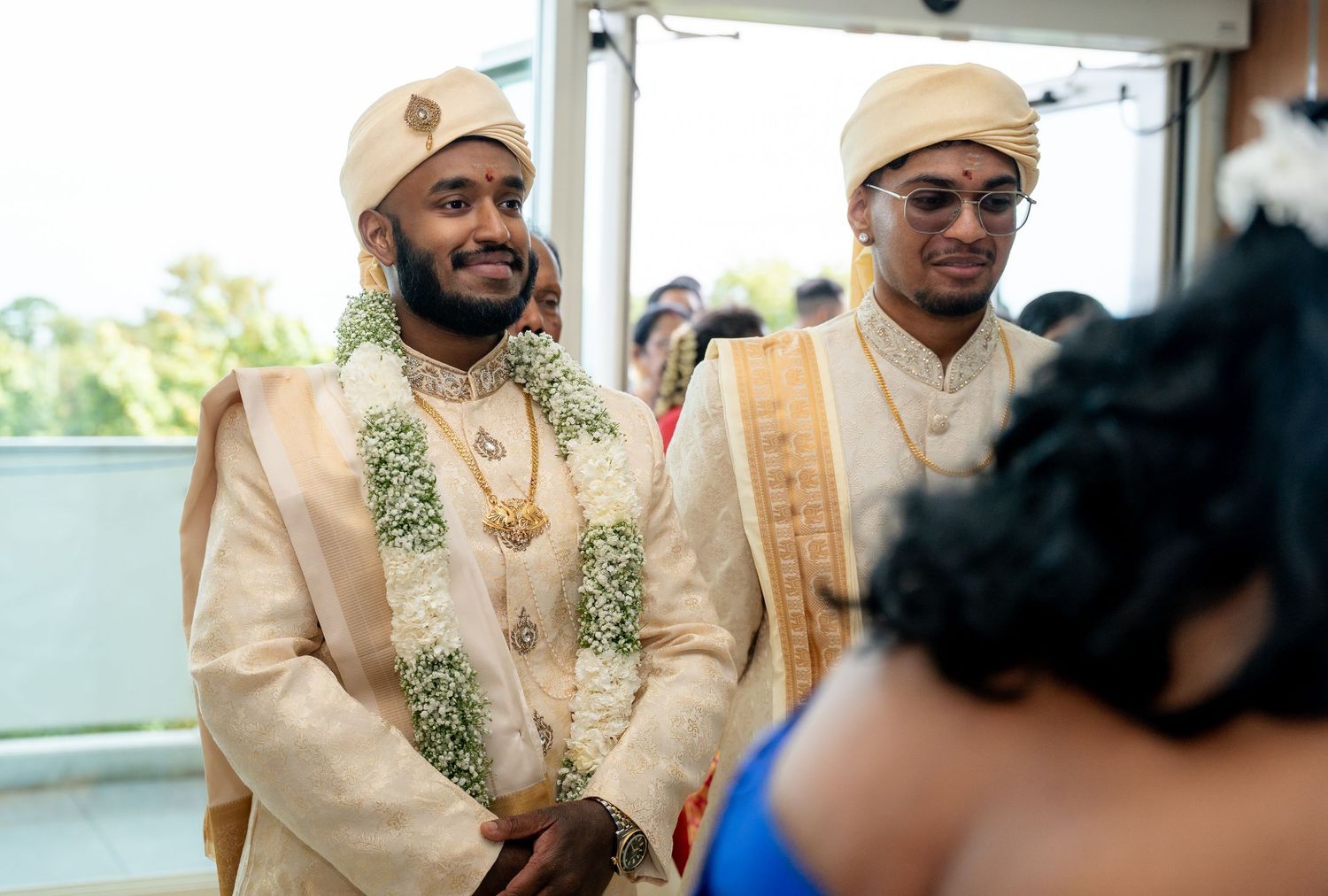 Two grooms in cream sherwanis and flower garlands smiling joyfully during a traditional Hindu wedding ceremony.