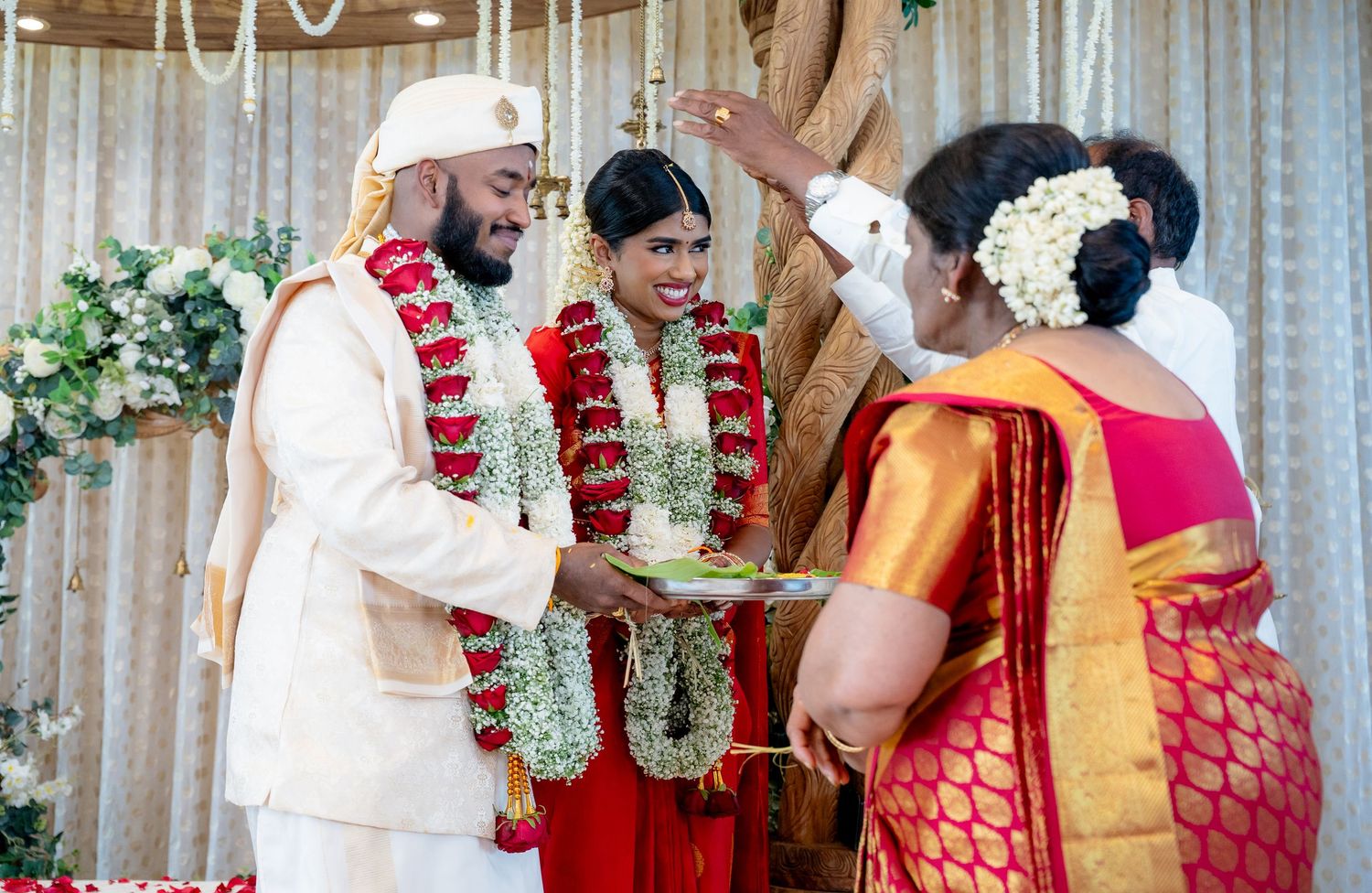 Bride and groom in red and white traditional attire receive blessings during South Indian wedding ritual ceremony.
