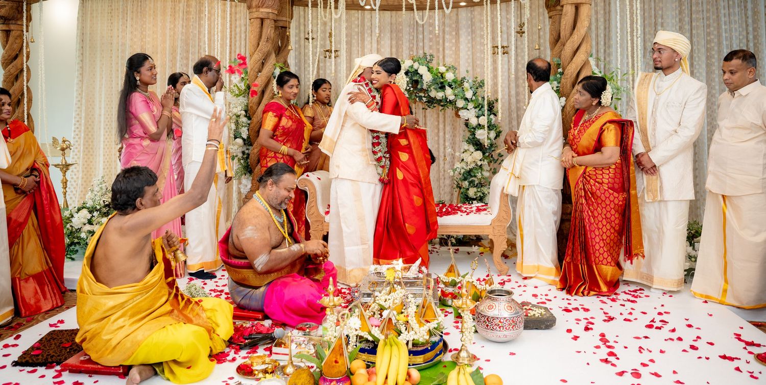 South Indian wedding couple exchange garlands on decorated mandap surrounded by family in traditional attire.