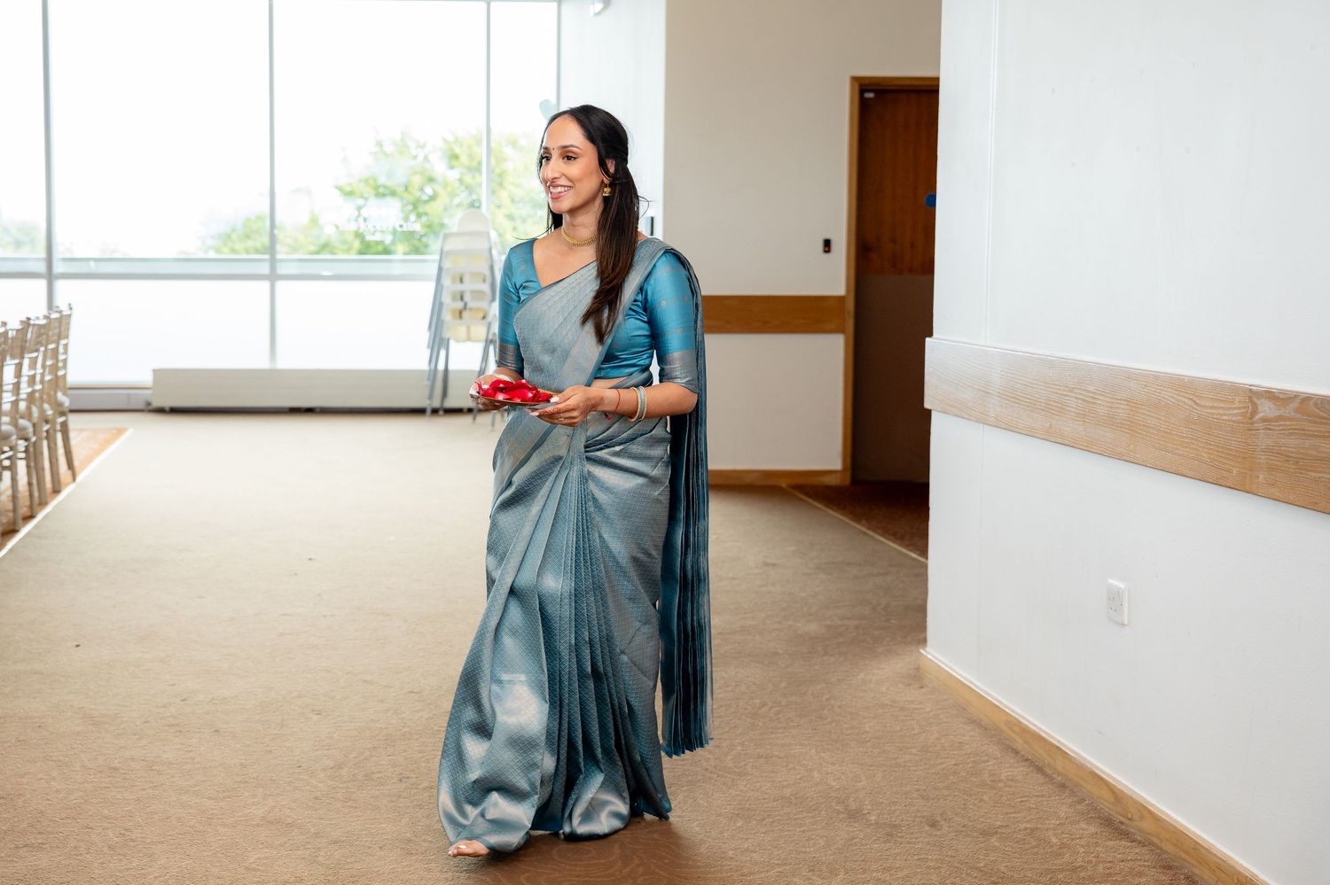 Elegant woman in teal silk saree carrying red tray walking through wedding venue corridor.