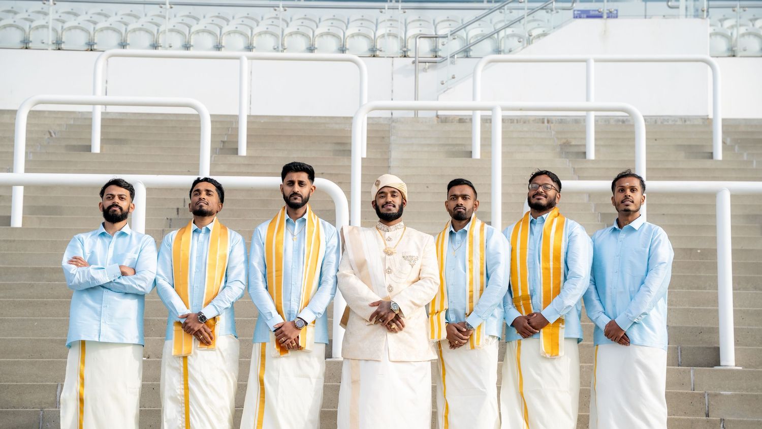 Seven men in traditional South Indian white and gold attire pose together in a stadium setting.