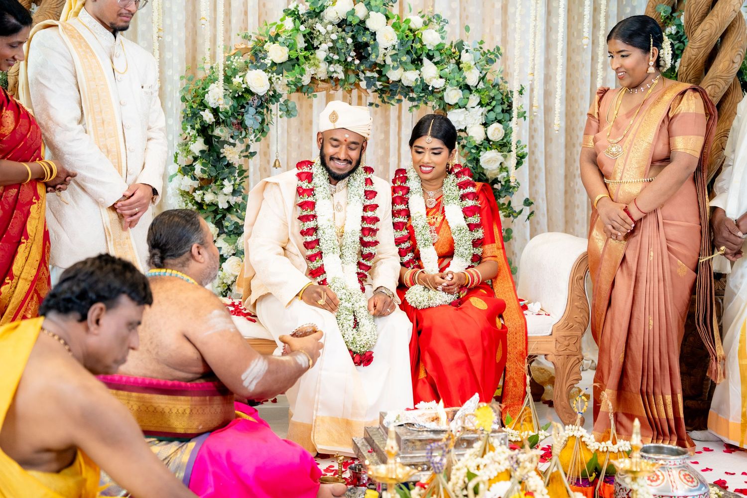 Bride and groom smile during traditional South Indian Hindu wedding ceremony with priest and family present.