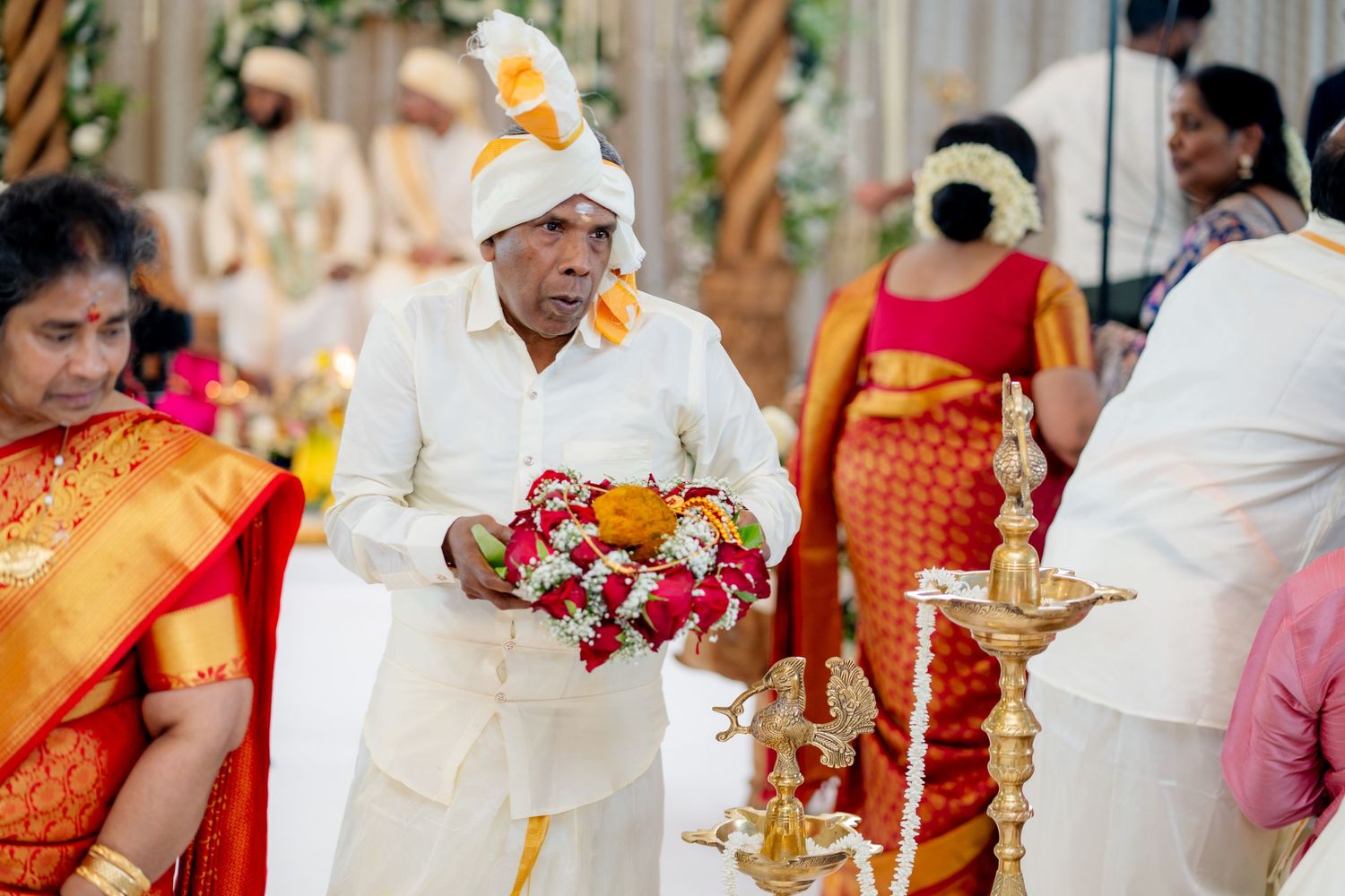 Elder man in white dhoti carries red and white flower garlands past oil lamp at South Indian wedding.