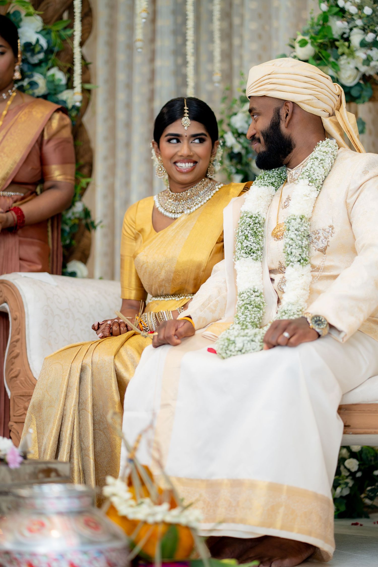 Smiling Tamil bride in gold Kanchipuram saree with jewelry and groom in cream sherwani at wedding mandap.