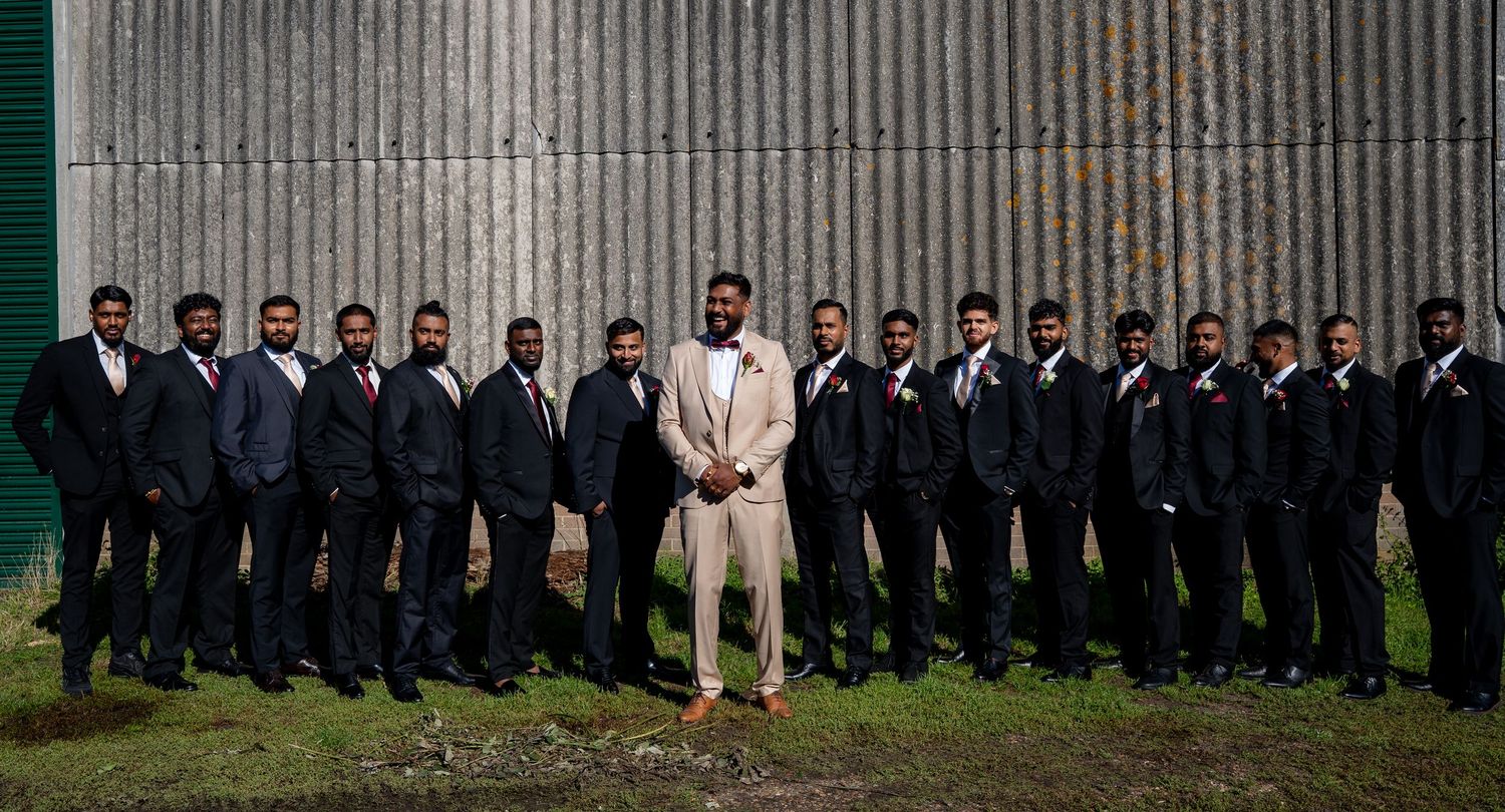 Groom in tan suit stands centered among groomsmen in dark suits against corrugated metal wall.