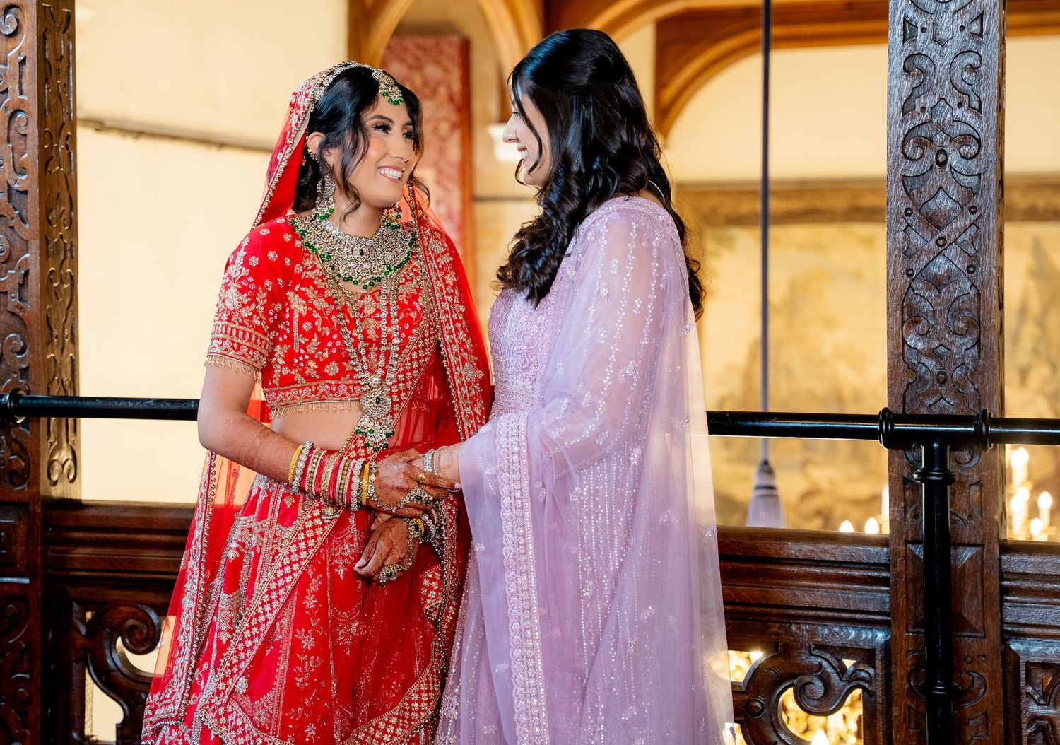 Indian bride in red lehenga sharing tender moment with bridesmaid in lavender outfit on balcony.
