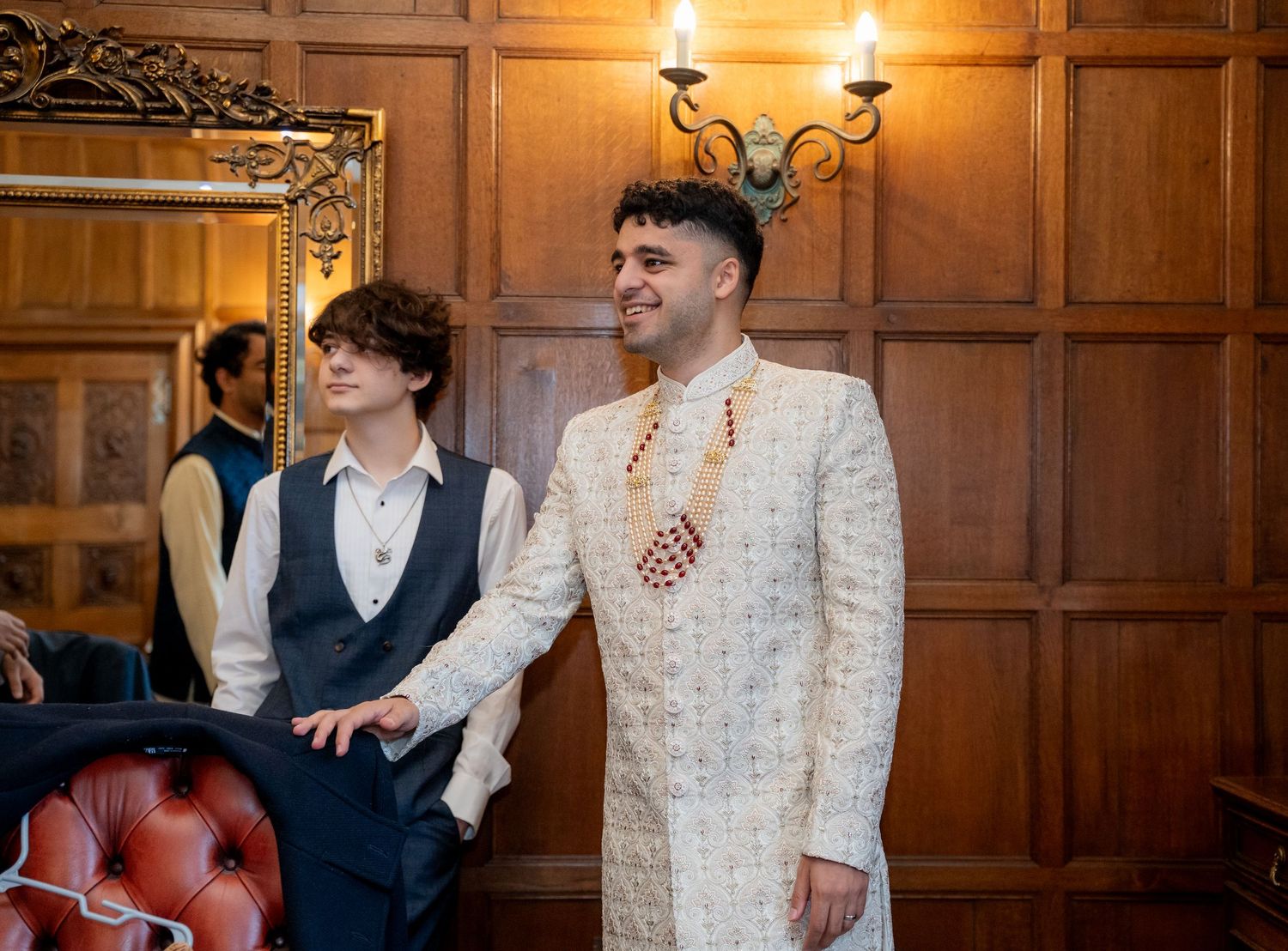 Groom in cream embroidered sherwani stands with guest in wood-paneled room at Indian wedding.