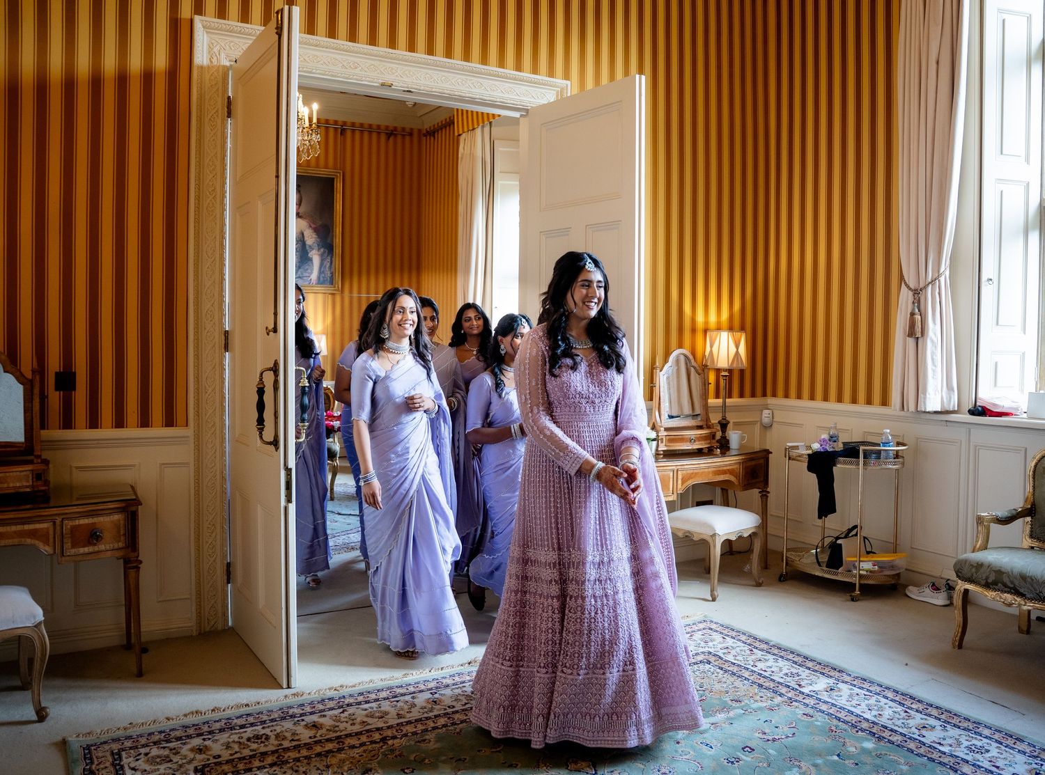 Bridesmaids in lavender gowns entering elegant yellow striped room.