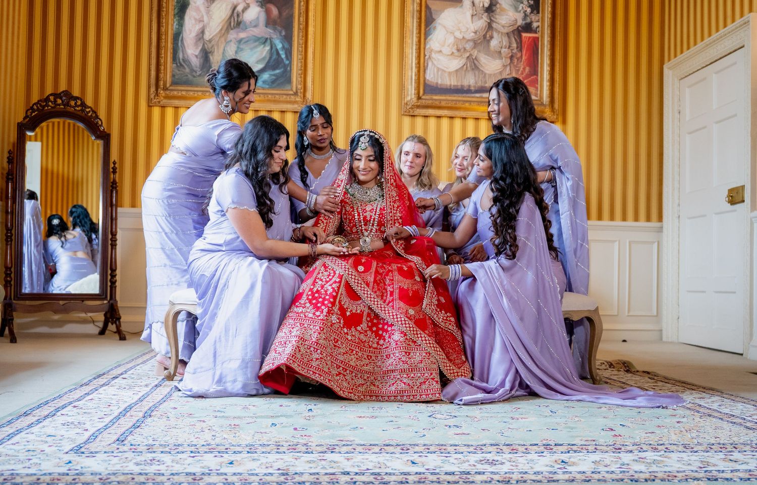 Indian bride in red lehenga surrounded by bridesmaids in lavender gowns during wedding preparations in elegant room.