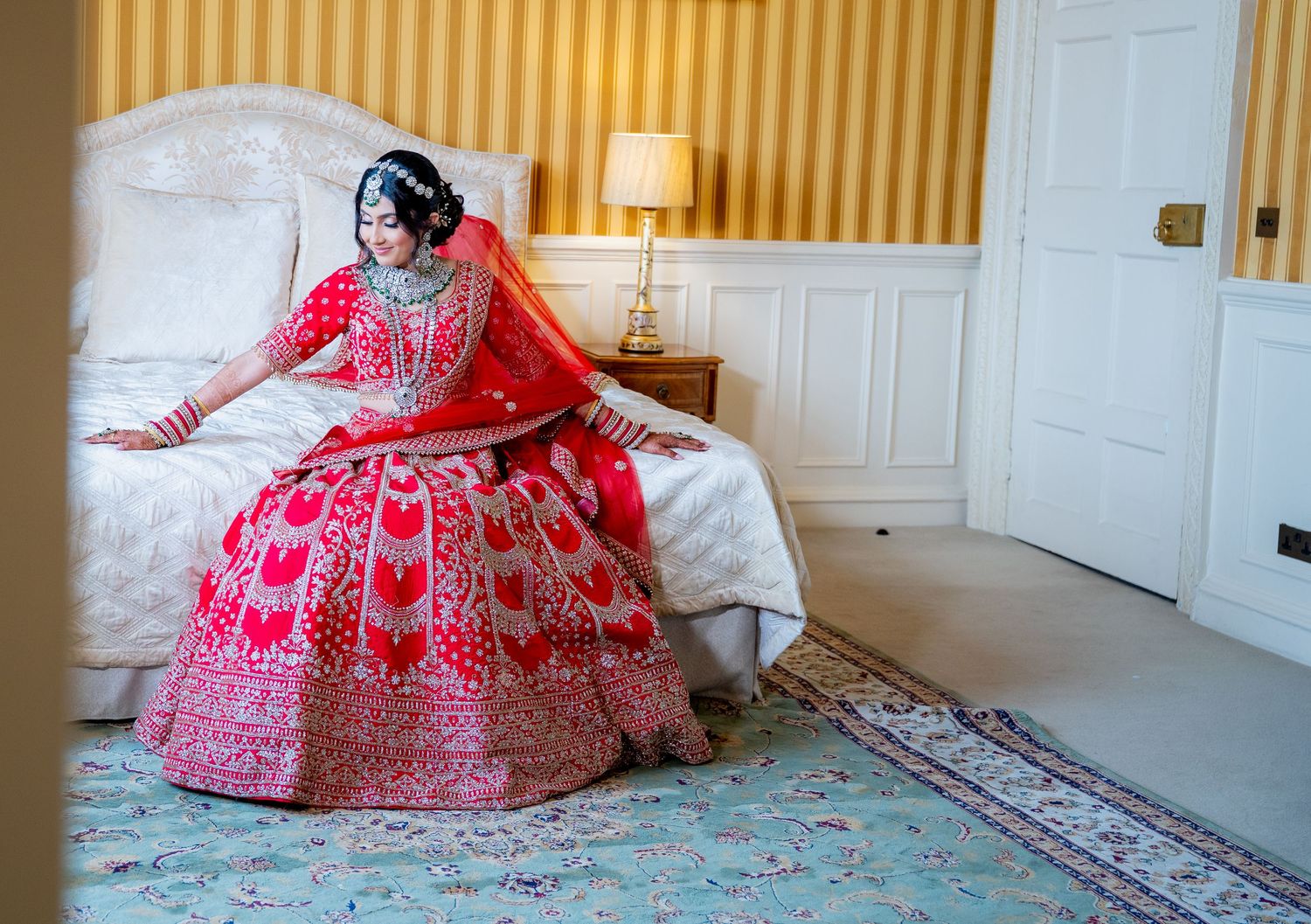 Indian bride in red bridal lehenga sitting on bed in yellow-striped elegant room.