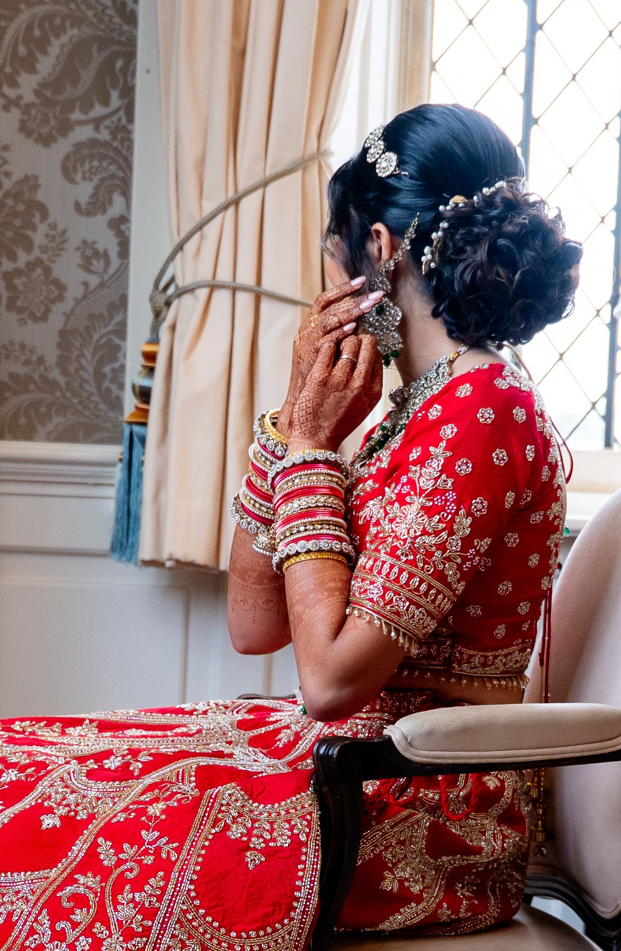 Indian bride in red lehenga adjusting earrings with mehndi hands and bangles by window light.