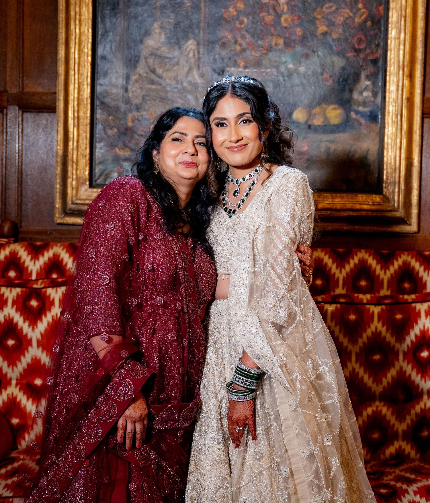 Bride in white lehenga with tiara poses with woman in red embroidered salwar at an elegant wedding venue.
