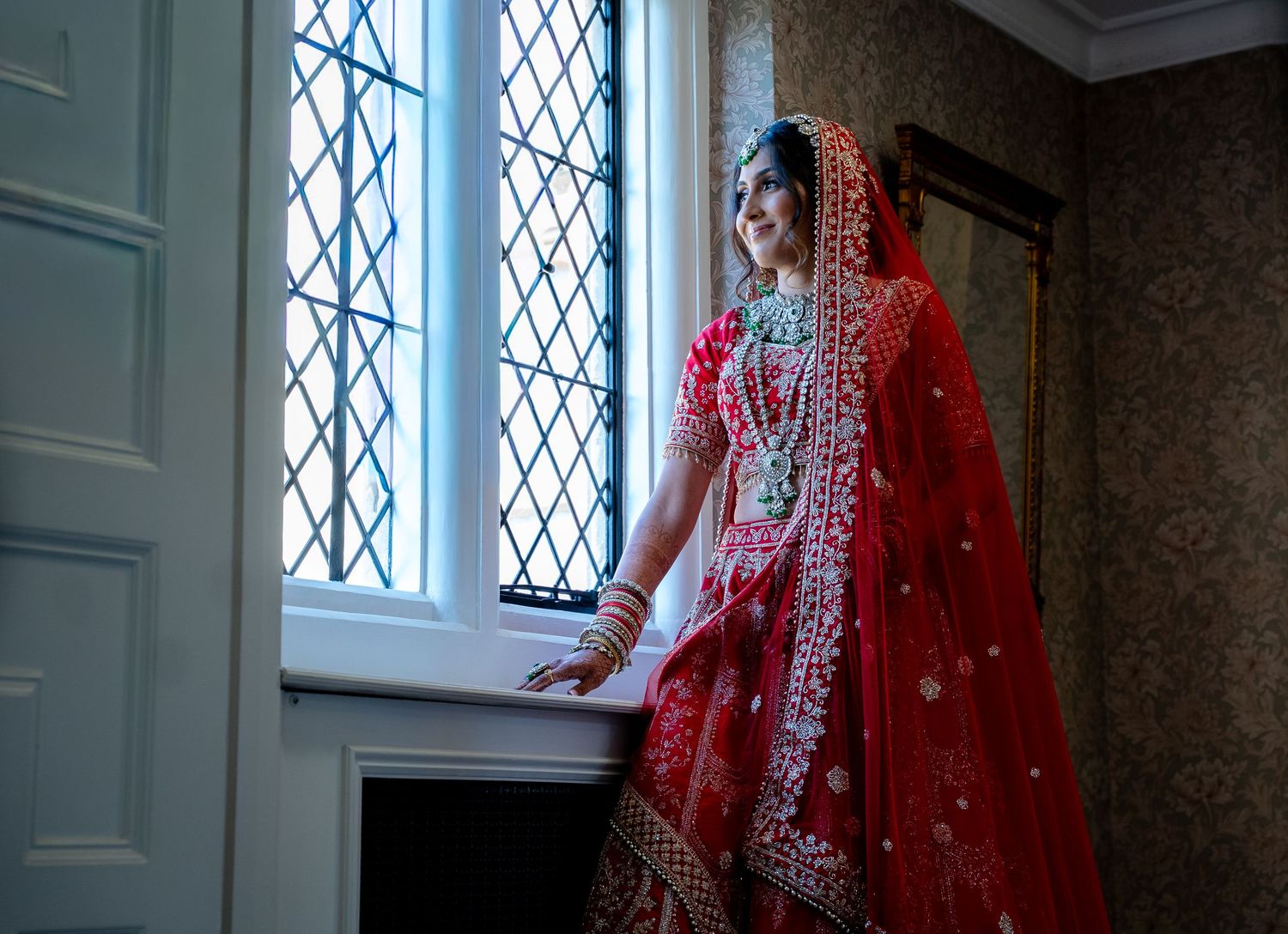 Indian bride in red bridal lehenga and dupatta standing by a latticed window.