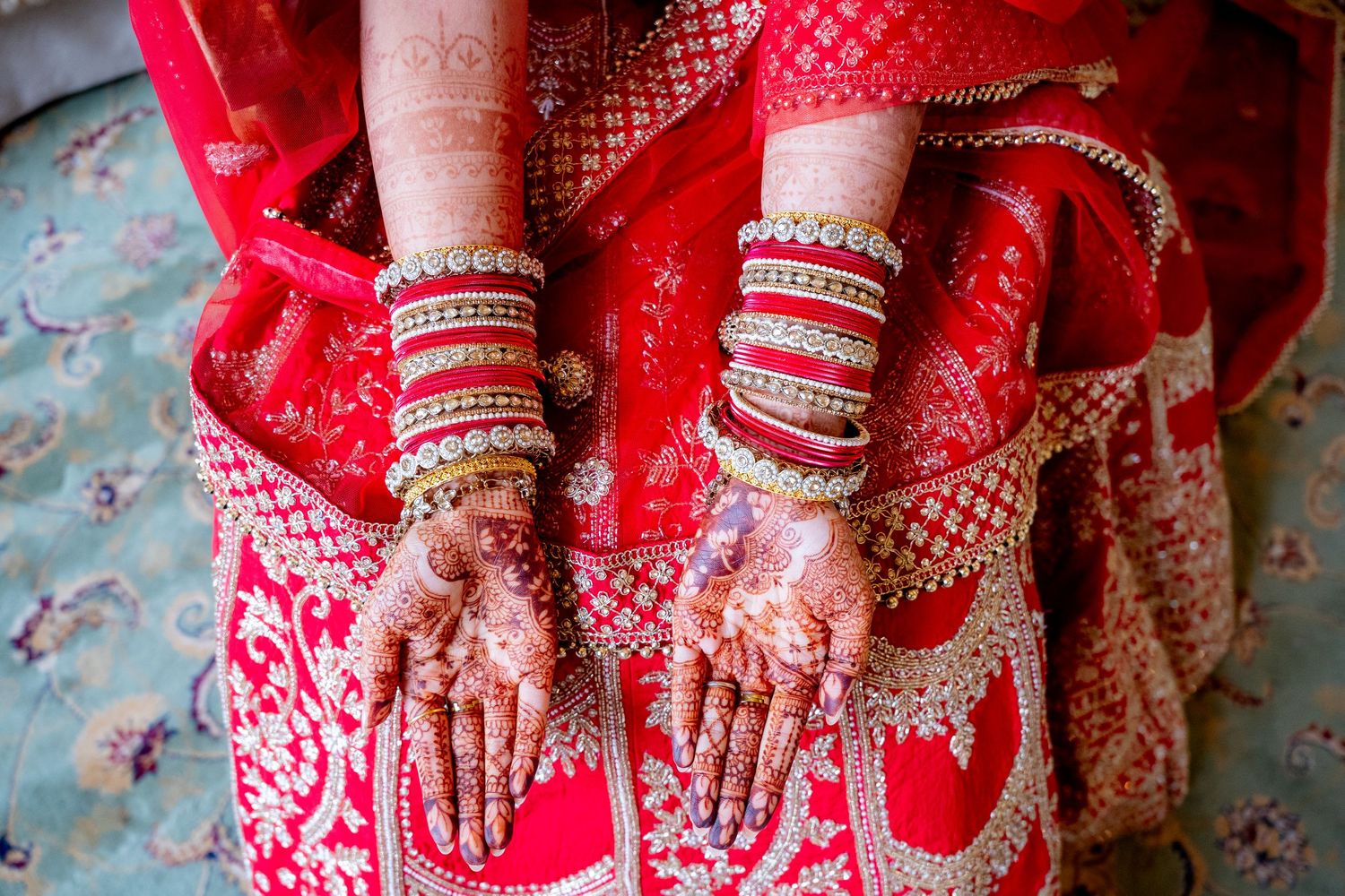 Close-up of Indian bride's hands adorned with intricate mehndi and gold bangles in red lehenga.