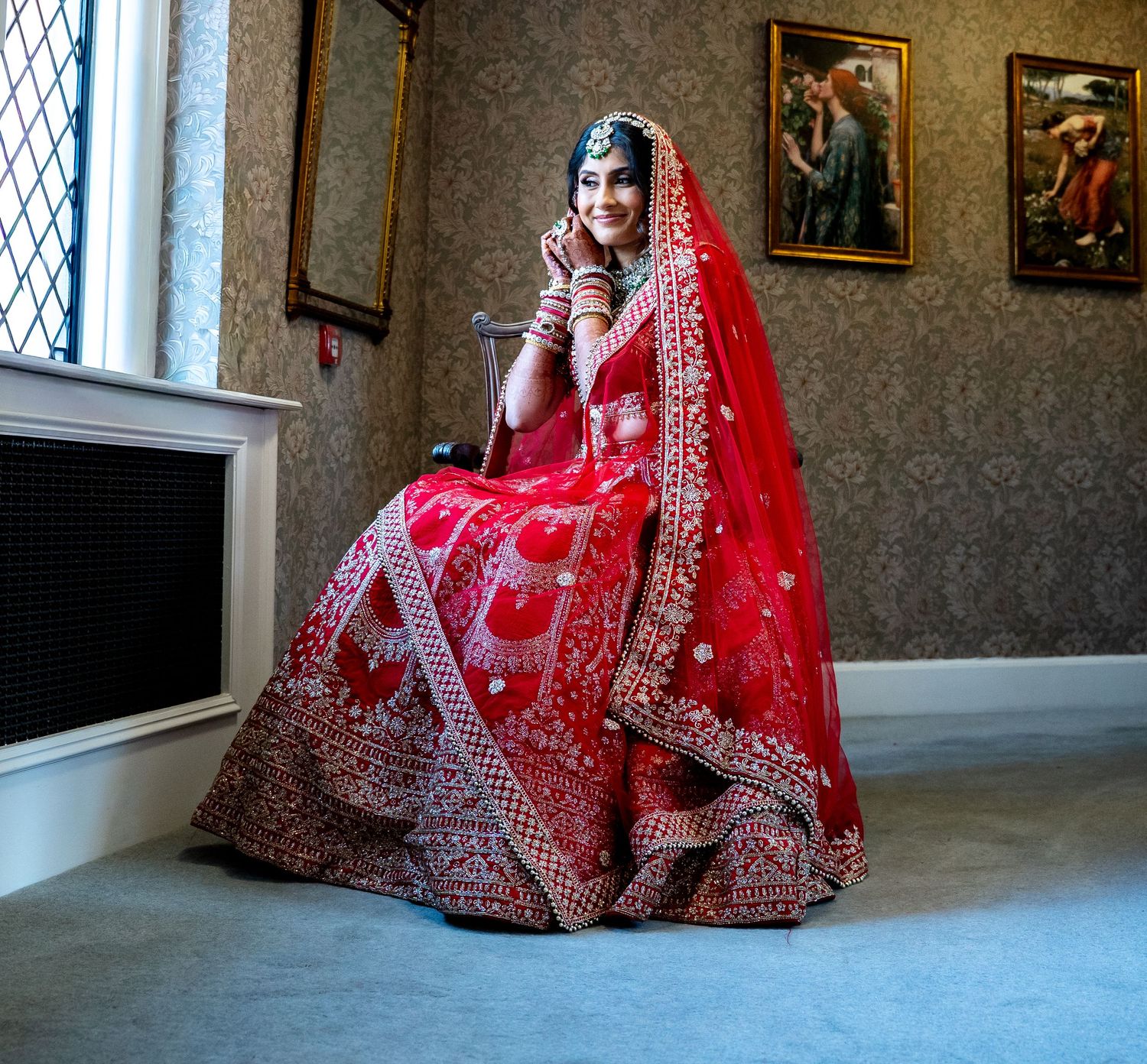 Indian bride in red lehenga sitting gracefully by window in elegant room.